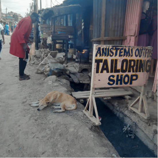 Tailoring shop sign sitting directly above an open drainage trench carrying stagnant sewage, reflecting daily business challenges amidst the climate crisis.