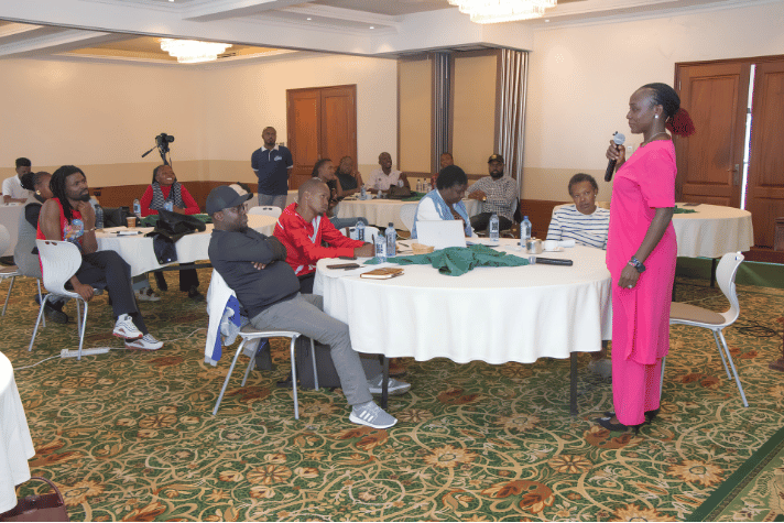 A facilitator in a pink outfit speaking into a microphone during an AMWIK capacity-building workshop for journalists seated around round tables.