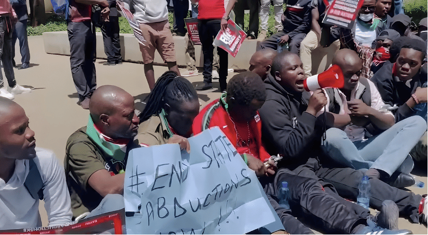 Activists sitting on the ground in a protest holding a banner reading "#END STATE ABDUCTIONS" with one demonstrator speaking through a red megaphoned horn.
