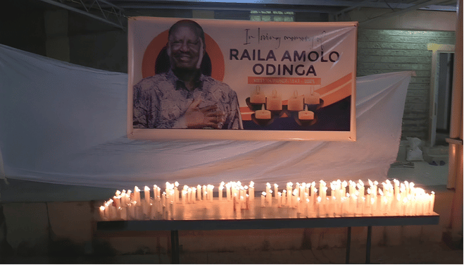 A memorial banner of the late Raila Odinga with rows of lit white candles arranged on a table in front of it during a night vigil.