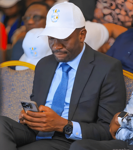 Edwin Sifuna wearing a white TEP cap and suit, looking down at his smartphone during a political event.