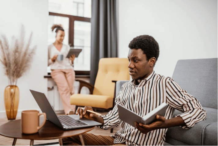 A young African man sitting in a modern room taking notes while working on a laptop, with a colleague in the background using a tablet, highlighting flexible remote jobs