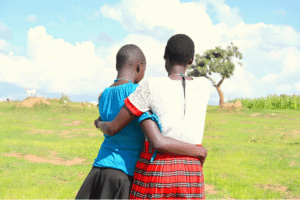 Two young girls embracing outdoors in a rural Kenyan field highlighting peer support to prevent child marriage