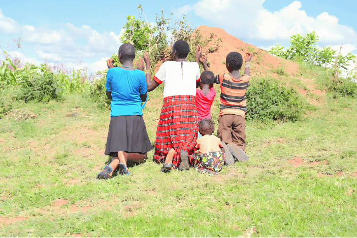A rural Kenyan family and children kneeling outdoors in prayer during a community intervention program against child marriage
